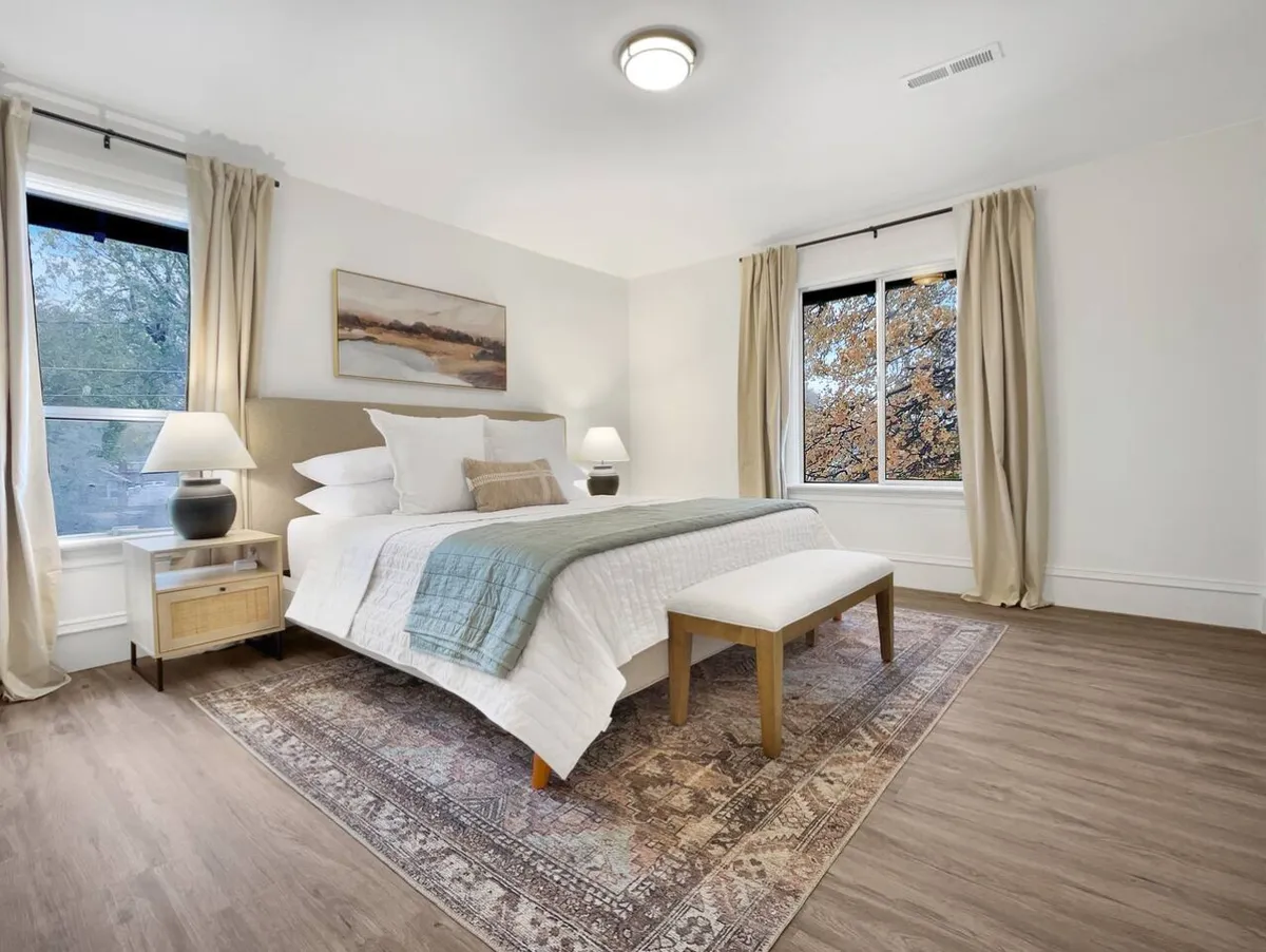 A cozy bedroom with a bed dressed in white linens, a large window allowing natural light to fill the space, decorative artwork on the wall, and a rug on the floor. The overall mood is tranquil and inviting.