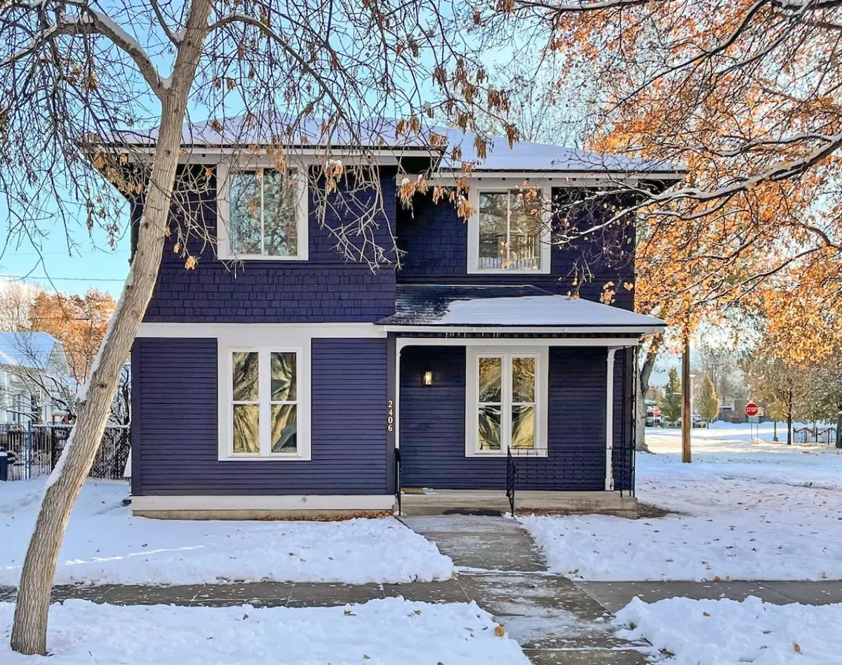 The image displays the exterior of a beautifully renovated purple single-family house in Baker City's Historic District. The snow-covered yard and bare trees evoke a cozy winter ambiance, while the inviting front porch hints at warm gatherings and relaxation. The scene suggests opportunities for guests to play in the snow, enjoy the peaceful neighborhood, and cozy up after outdoor adventures.