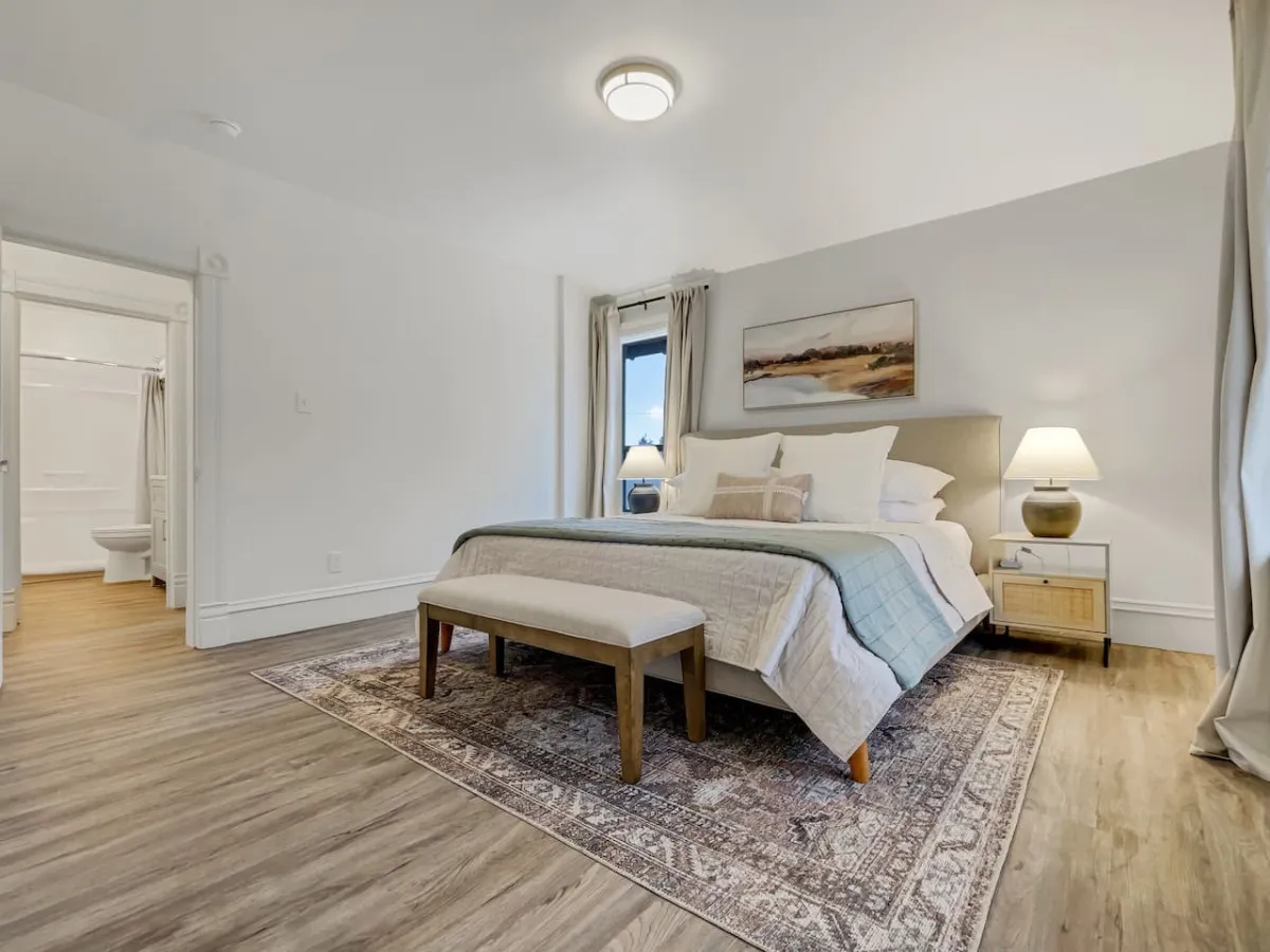 A king-sized bed with soft linens, a nightstand and lamp, a bench at the foot of the bed, and an area rug beneath. The room is bathed in natural light from the window, and the decor is calm and serene.
