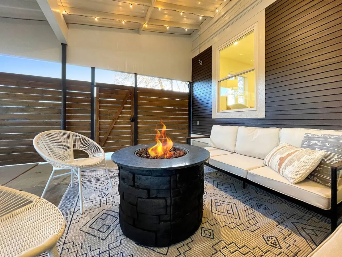 An exterior shot of a stylish patio area with a fire pit at its center, surrounded by a comfy outdoor couch, chairs, and a decorative rug. The setting is ideal for group relaxation or lively gatherings, blending comfort with a touch of modern style.