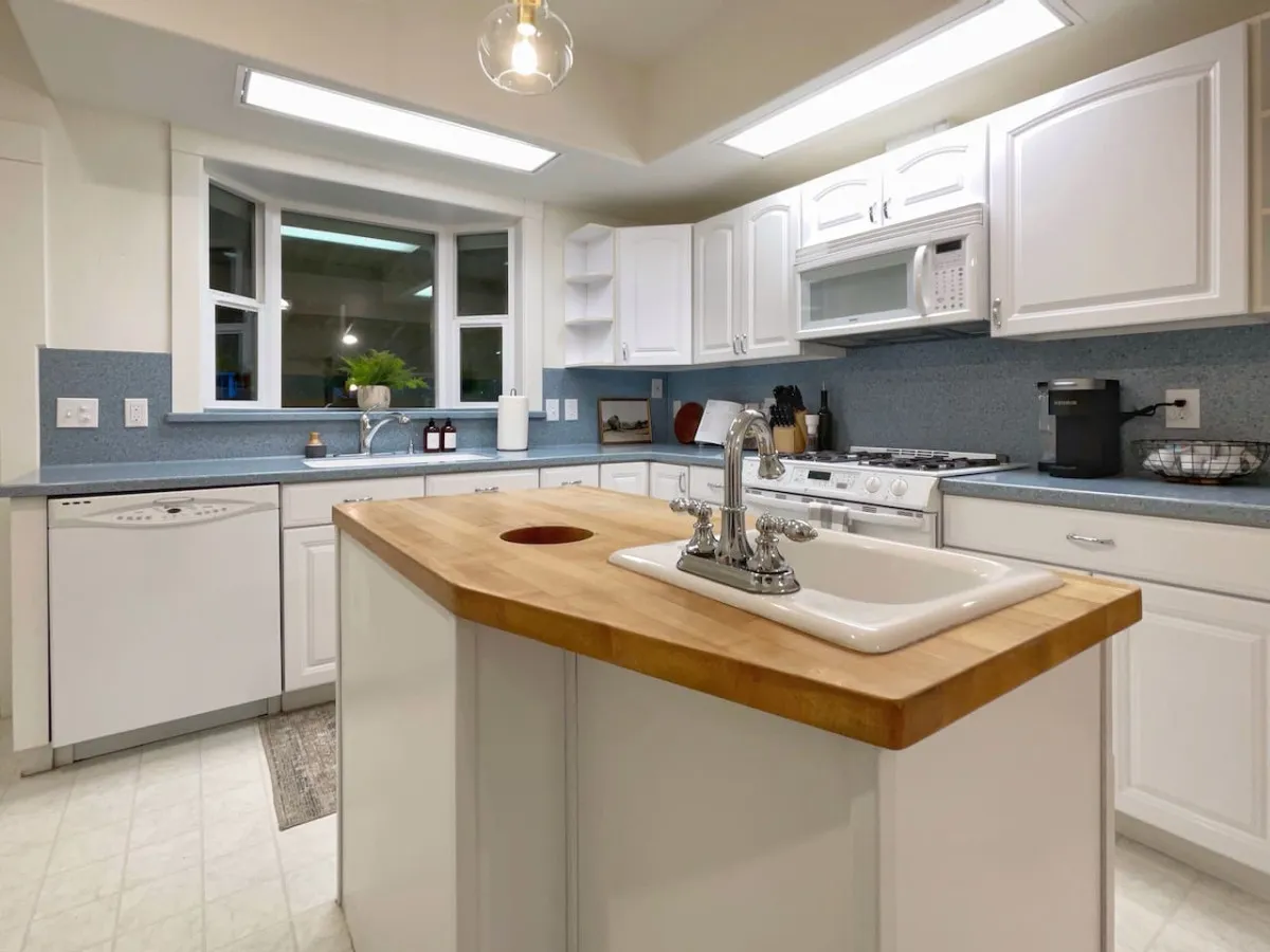 This image features a bright and inviting kitchen with a central island that includes a sink, a window bringing in natural light, and a view of the stove and microwave area. The counters are spacious and modern appliances are visible, creating an atmosphere that encourages cooking and socializing.