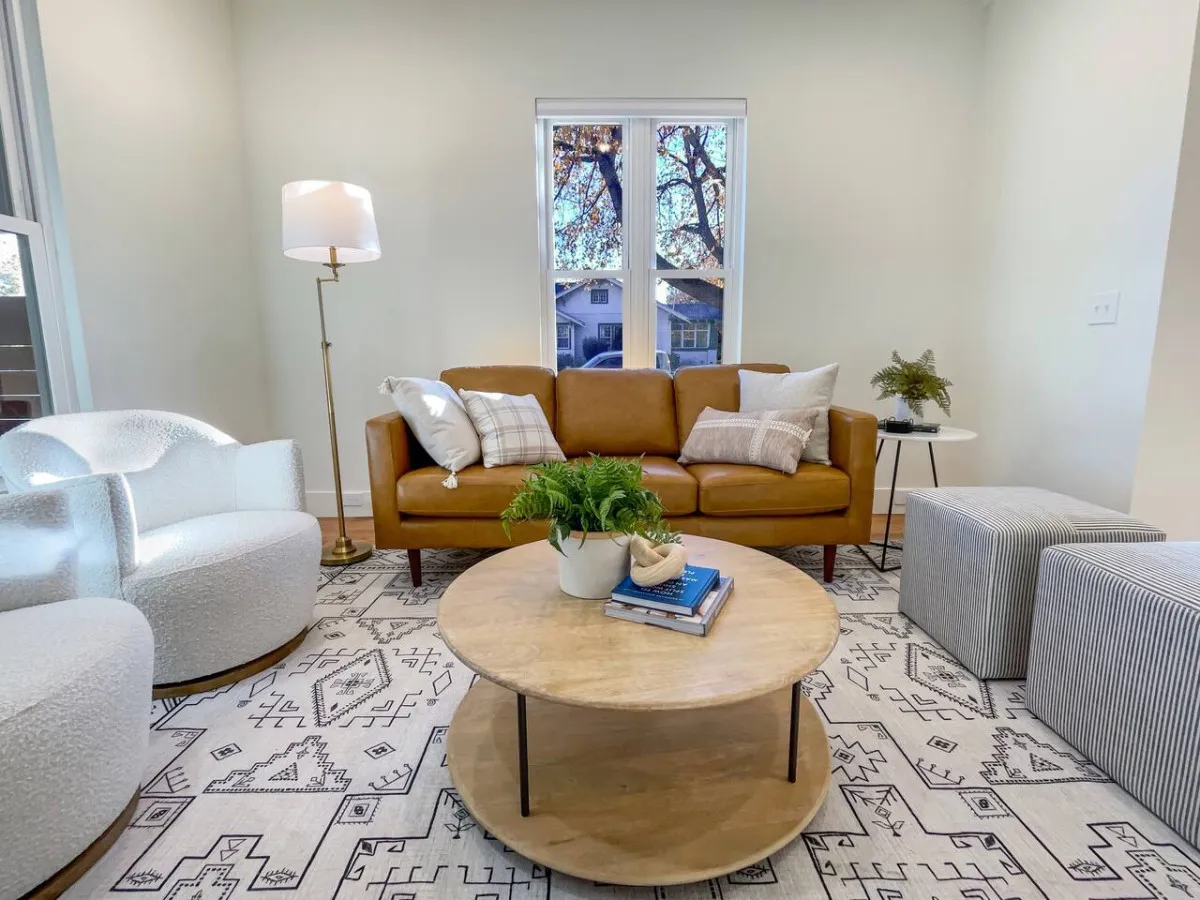 A cozy living area featuring a brown leather sofa, round coffee table, and stylish armchairs arranged by a large window. Natural light fills the room, highlighting the plush furnishings and creating an inviting, sunlit atmosphere perfect for relaxing or socializing.