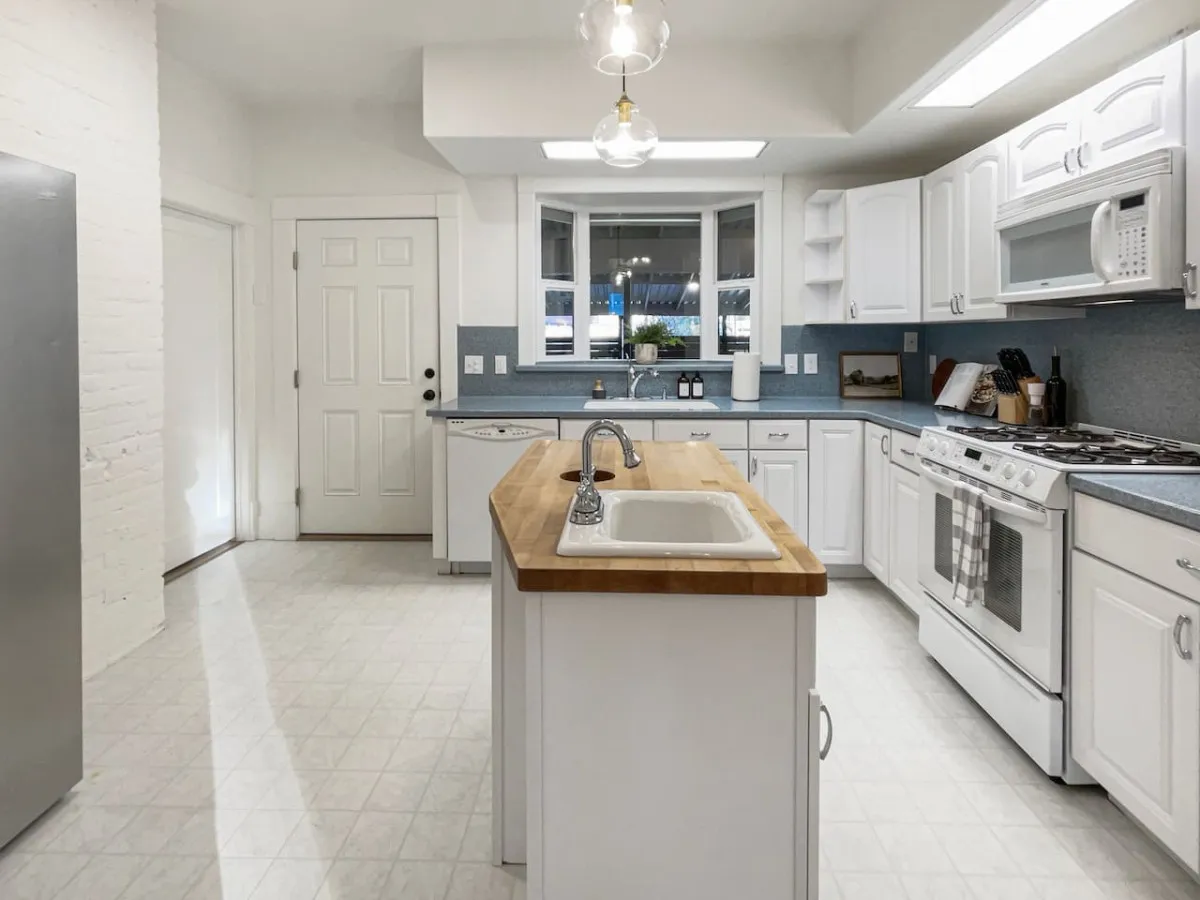 A warmly lit kitchen scene showcasing the kitchen island, a sink, stove, and window. The overall ambiance is welcoming, with plenty of space for multiple people to cook or gather, reflecting a hub for family gatherings and entertaining.