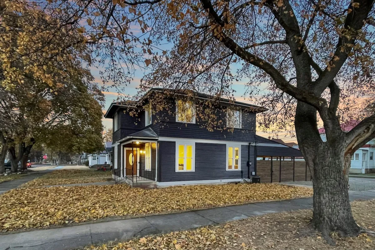 A two-story house with warmly lit windows is surrounded by mature trees and fallen autumn leaves. The tranquil exterior exudes a sense of welcome and comfort, with hints of cozy evenings awaiting within.