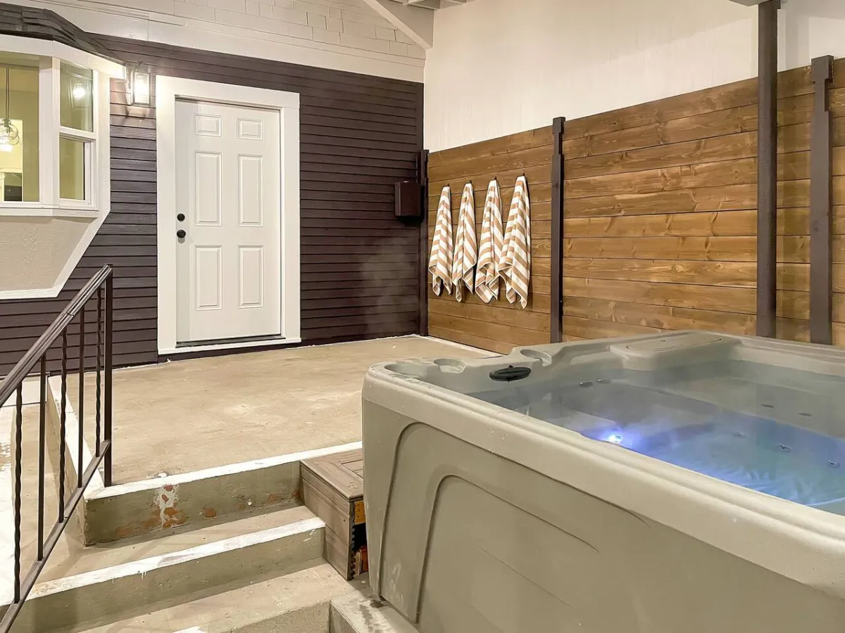 A tranquil exterior view of the home's patio area featuring a bubbling hot tub. The front door is visible in the background, with plush towels nearby, inviting guests to unwind. The scene is set at dusk, suggesting a perfect setting for stargazing and relaxing after an active day.