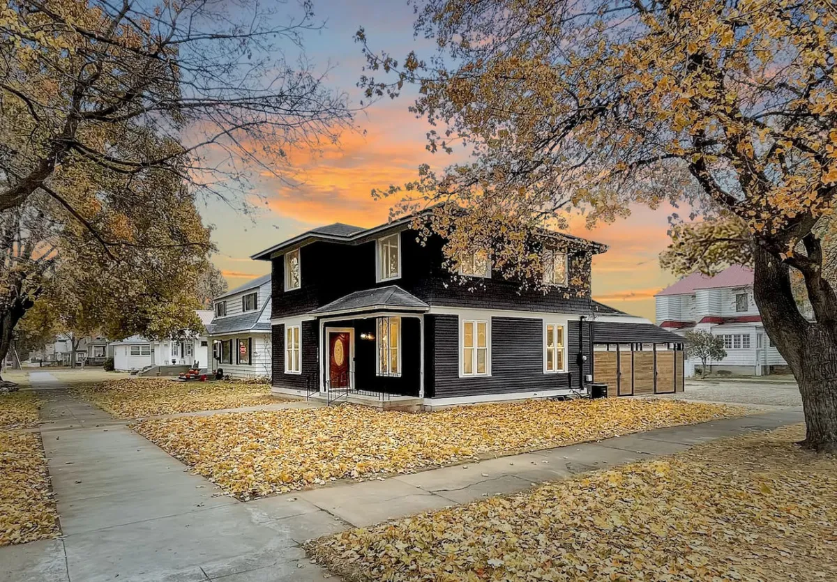 A view of the house exterior at sunset, featuring its black façade, welcoming front door, fallen autumn leaves, and softly glowing windows, evoking a sense of arrival and homecoming. The setting is cozy and inviting, hinting at the warmth and hospitality inside.
